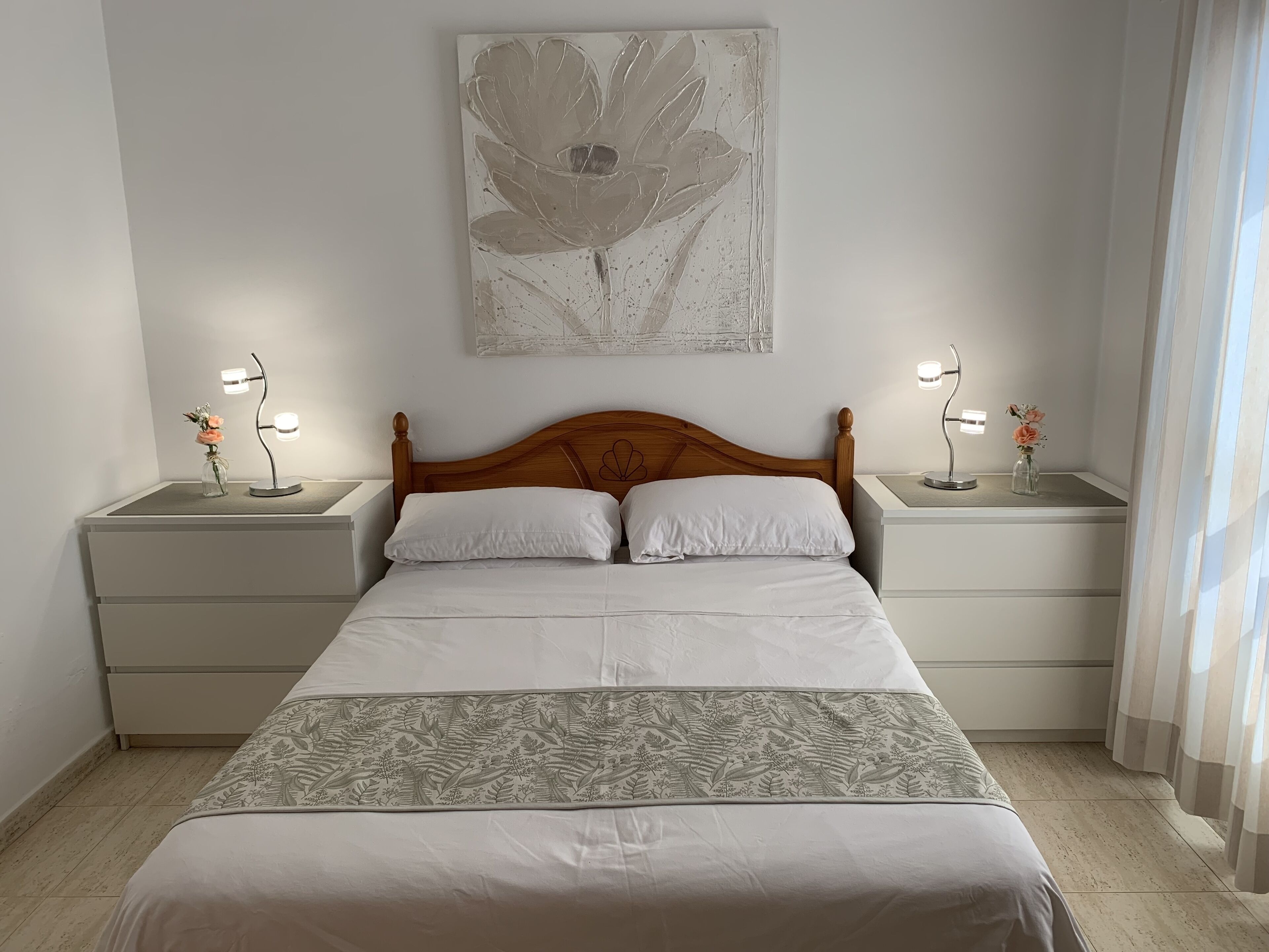 A neatly arranged bedroom with a wooden headboard, white bedding, two bedside tables with lamps, and a floral artwork on the wall.