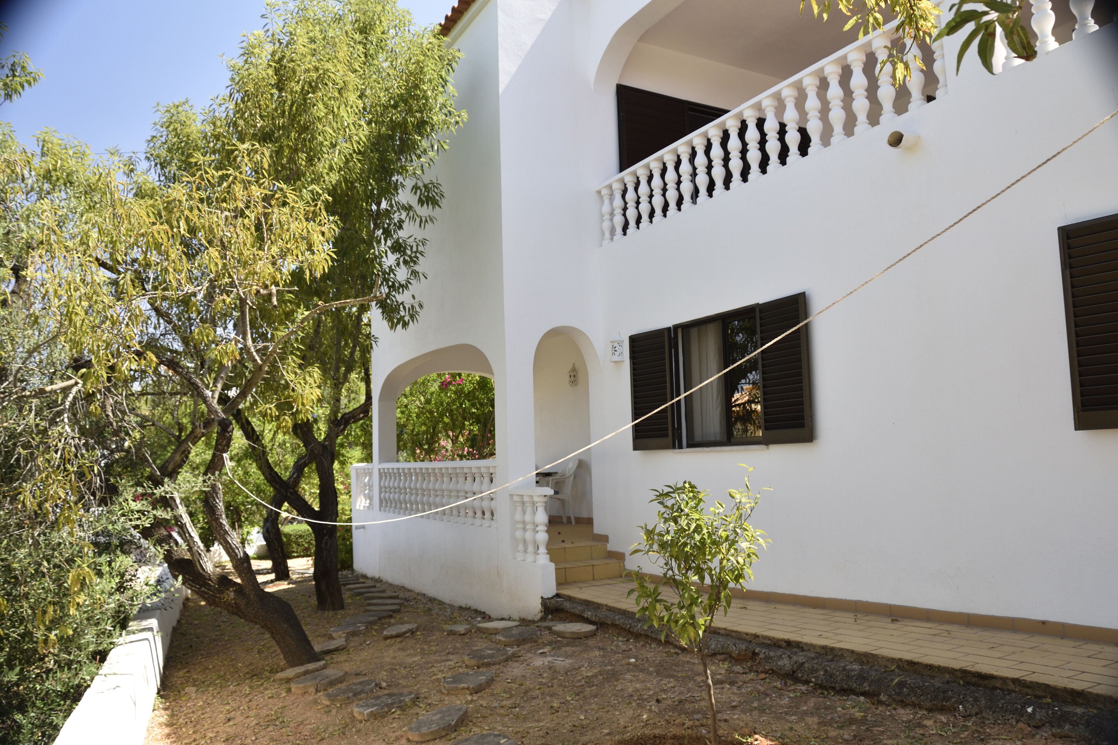 A white building with a balcony, a small garden, and a tree.