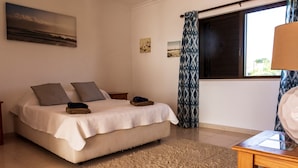 A bedroom with a bed, a wooden nightstand, a small table, a window with curtains, and a wall with a picture. - Large Villa with own Pool, 4 Bedrooms, Family Friendly, Shaded Pavillion. (Vale Covo, Loule)