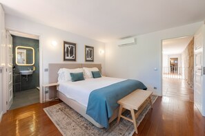 A bedroom with a bed, bedside table, bench, wall-mounted air conditioner, and a bathroom with a sink and mirror.