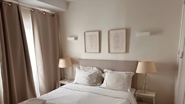 Una cama bien hecha con sábanas blancas y dos almohadas, dos lámparas de noche y dos marcos con textos en la pared.