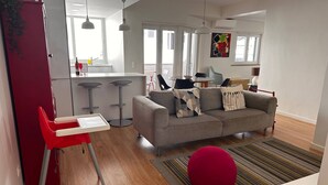 Una sala moderna con un sofá gris, una silla roja y un reposapiés rojo. Cuenta con una zona de cocina con un refrigerador, taburetes de bar y una mesa de comedor.