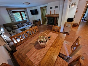 A cozy living room with a sofa, a wooden dining table with chairs, a fireplace, and a television mounted on the wall.