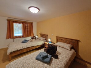 A room with two wooden beds, a window with curtains, a radiator, a lamp, and a teddy bear on one bed.