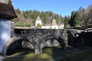 Un puente de piedra con arcos, un edificio blanco de techo cónico y un paisaje montañoso cubierto de bosque al fondo.