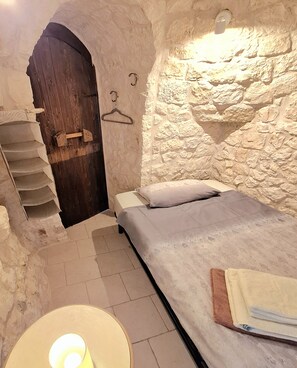 A stone-walled room with a bed, a wooden door, a staircase, and a hanging light fixture.