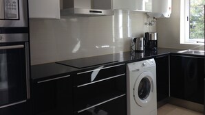 Une cuisine moderne équipée d’appareils électroménagers en acier inoxydable, d’un four encastré, d’un lave-linge et d’une fenêtre laissant entrer la lumière naturelle.