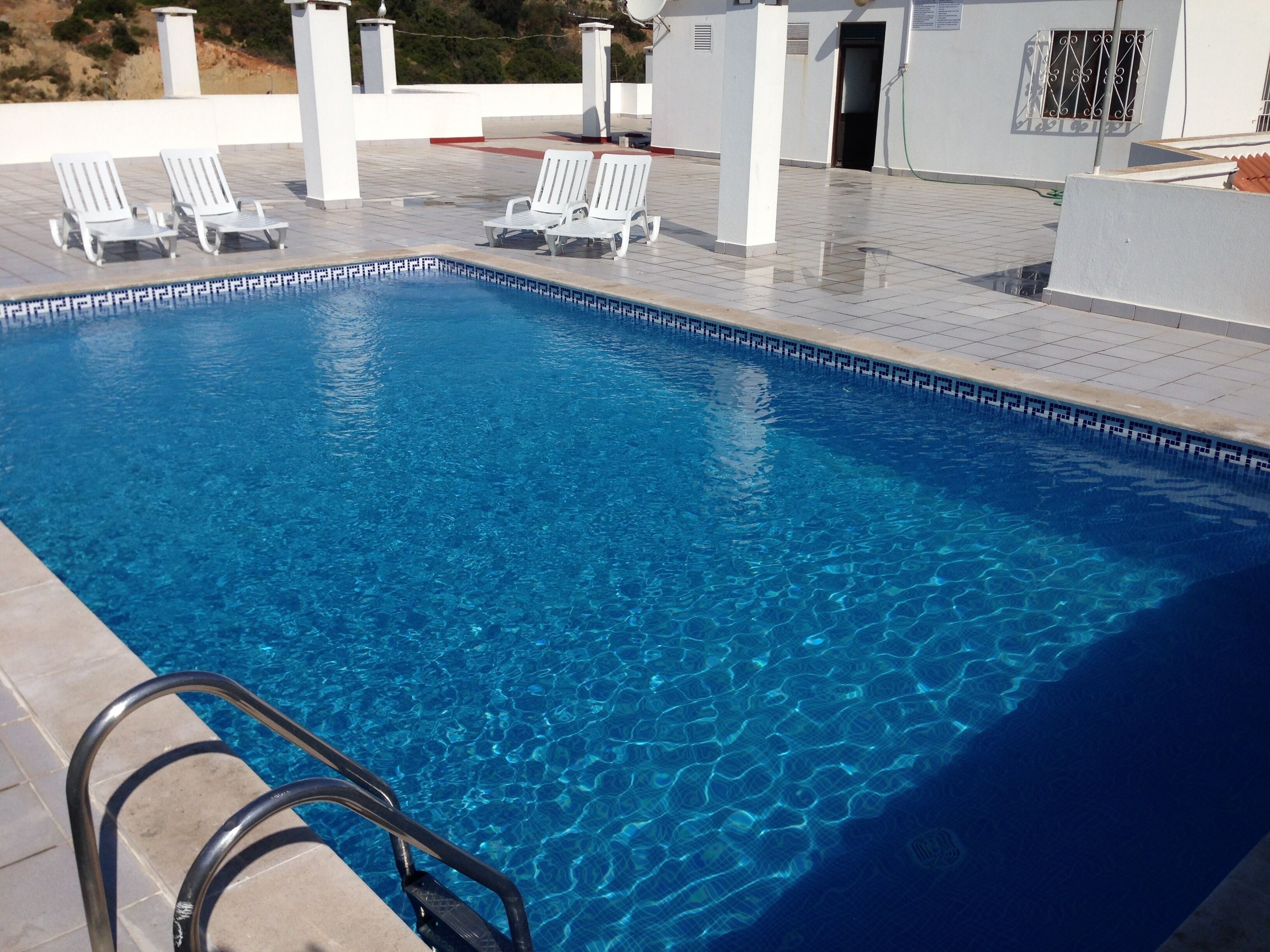 Una piscina rectangular con agua azul cristalina, rodeada de sillas de descanso blancas y una escalera metálica.