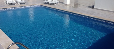 Una piscina rectangular con agua azul cristalina, rodeada de sillas de descanso blancas y una escalera metálica.