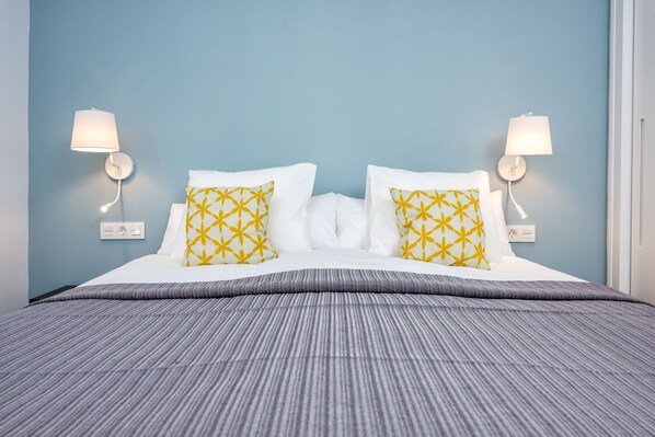 A neatly made bed with white and yellow patterned pillows, two wall-mounted lamps, and a grey striped bedspread.