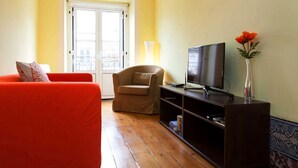 A living room with a red sofa, a beige armchair, a TV on a wooden stand, and a vase with flowers. - Apartment Baixa/Chiado view Castle (Lisboa)