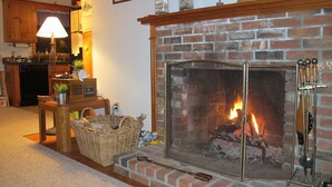 Una sala de estar acogedora con chimenea de ladrillo, una mesa de madera con lámpara, y una cocina al fondo.