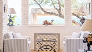 Une pièce avec deux canapés à rayures, une cheminée, une table avec une lampe et une grande fenêtre donnant sur un arbre et une peinture représentant un chameau.