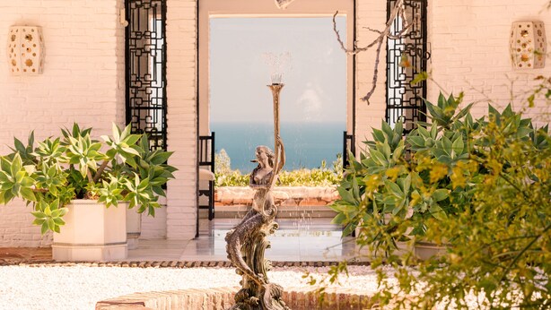 Une statue d’une femme tenant une torche, entourée de plantes en pot, avec une vue sur la mer en arrière-plan.