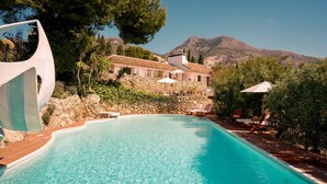 Une piscine bordée d’un mur en pierre, avec des chaises longues et une montagne en arrière-plan.