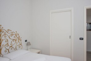 Una camera da letto con un letto bianco, un comodino bianco con una lampada e una porta bianca.