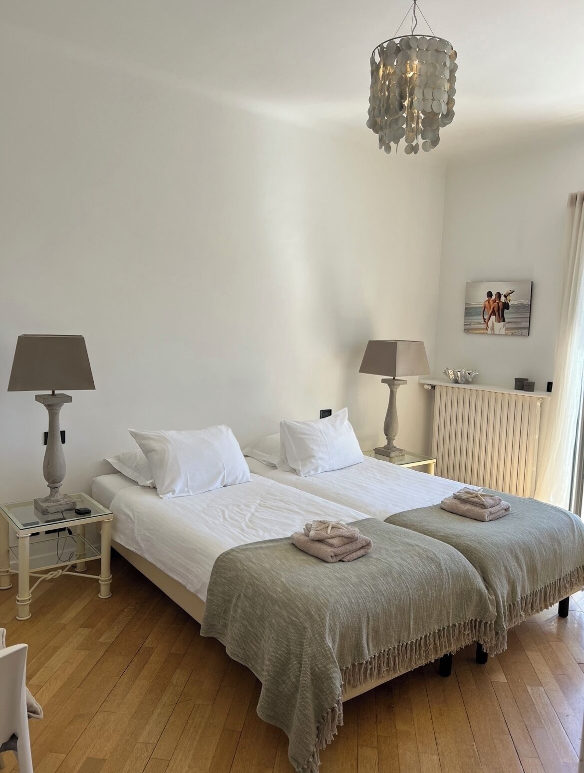 Uma cama de casal com roupa de cama branca, duas mesas de cabeceira com abajures, um candelabro e um quadro na parede.