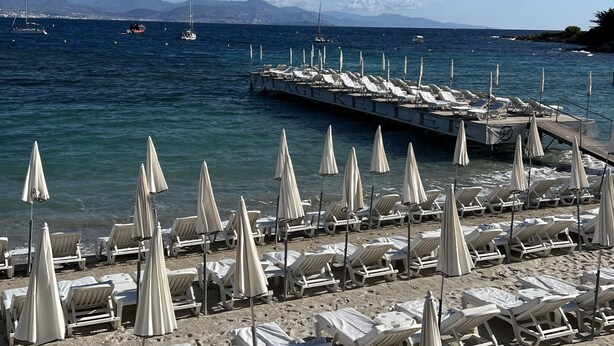 A beach with rows of sun loungers and umbrellas, a pier extending into the sea, and boats in the background.