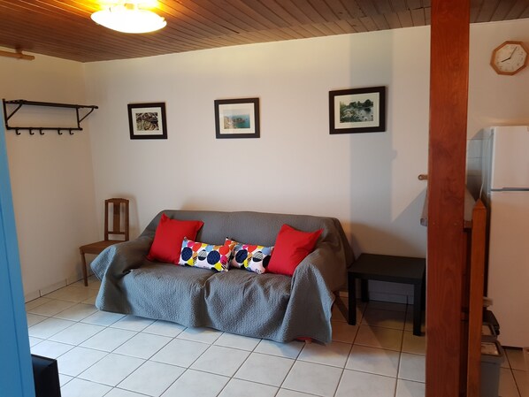 A cozy living room with a grey sofa, red and multicolored pillows, a wooden chair, and a small table. There are three framed pictures on the wall and a clock. - Champcervon: house / villa - Champcervon (Champcervon)