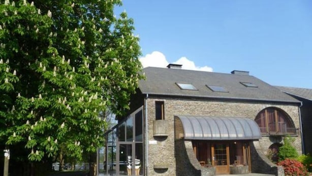 Un edificio de piedra con un amplio ventanal, un acceso cubierto y un árbol frente a él.
