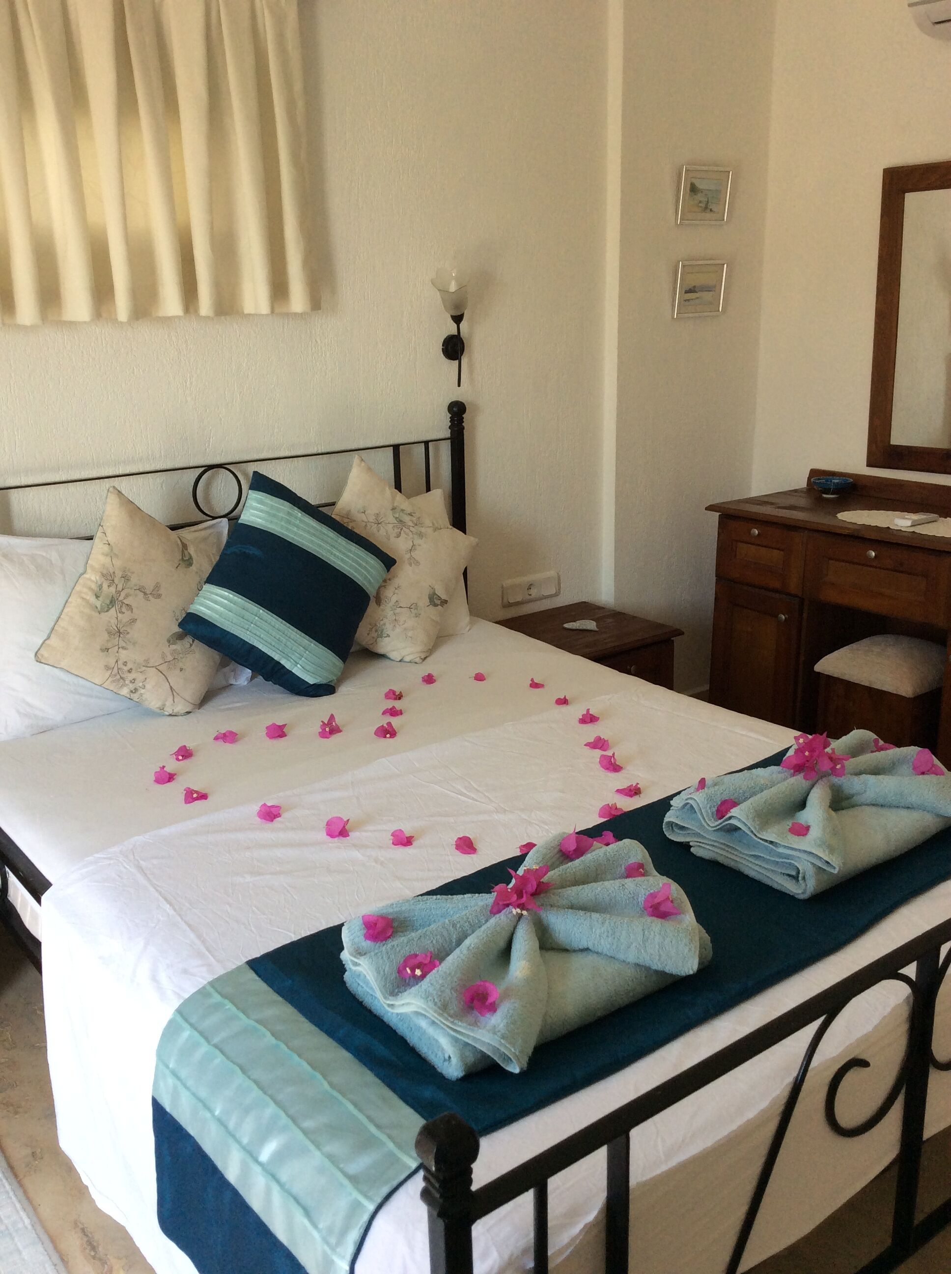 Un lit bien fait, agrémenté d’un coussin à motifs floraux, d’un coussin à rayures bleues et vertes, et de pétales de fleurs roses éparpillés sur le drap. Une serviette pliée, ornée de fleurs roses, est posée sur le bord du lit.