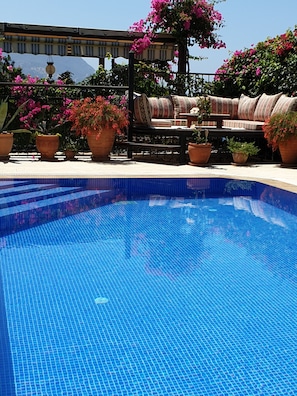 A swimming pool with tiled flooring, surrounded by potted plants and outdoor furniture.