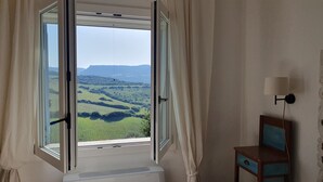 A room with a large window, a radiator, a small table with a lamp, and a view of green hills. - House in the countryside with wonderful views Asinara Gulf and Castelsardo (Castelsardo)