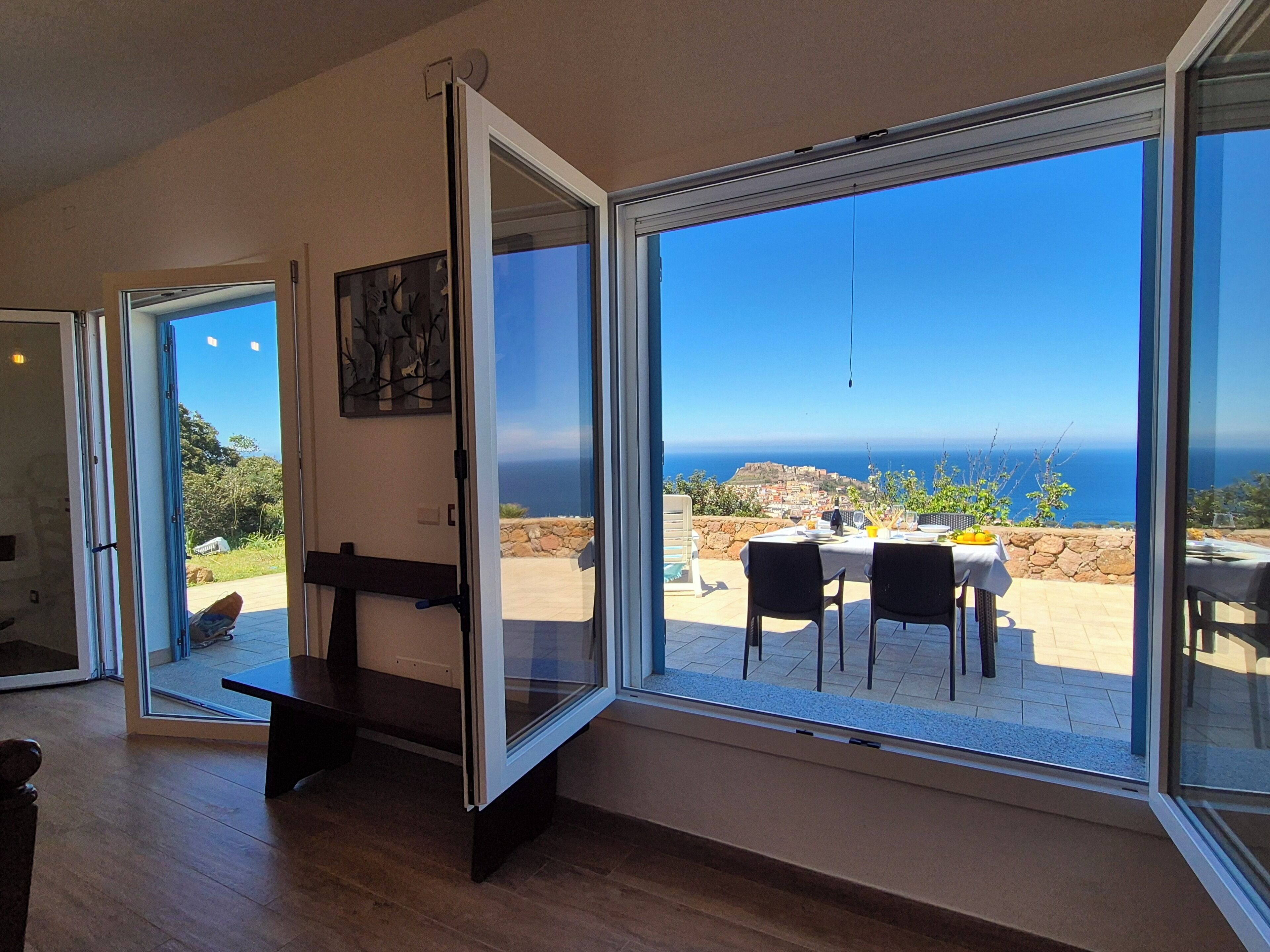 A room with a large sliding glass door leading to an outdoor patio with a table and chairs, a stone wall, and a view of the ocean.