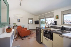 A compact kitchen with a refrigerator, stove, and sink, a dining area with orange chairs, and a large window providing a view of buildings outside.
