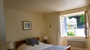 A bedroom with a bed, a bedside table, a lamp, a window with curtains, and a radiator. - Fisherman's Cottage with WiFi and views to the harbour. (Mortagne-Sur-Gironde)