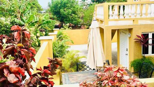 Ein gelbes Gebäude mit Balkon, umgeben von üppigem Grün und einer Terrasse mit Gartenmöbeln.