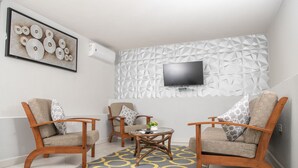 Ein modernes Wohnzimmer mit zwei Holzsesseln, einem Glastisch und einem gemusterten Teppich. Ein Flachbildfernseher ist an einer strukturierten Wand angebracht, und ein gerahmtes Kunstwerk hängt über den Sesseln.