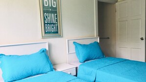 Ein Schlafzimmer mit zwei Betten, jeweils mit einer blauen gesteppten Bettdecke und passenden Kissen. Dazwischen steht ein weißer Nachttisch, und an der Wand hängt ein gerahmtes Schild mit der Aufschrift 'BIG SHINE BRIGHT'.