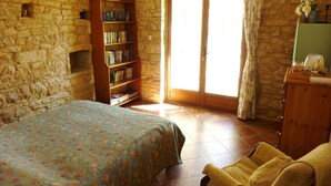 A bedroom with a stone wall, a wooden bookshelf, a bed with a floral bedspread, a yellow armchair, and a sliding glass door. - Beautiful retreat With Panoramic Countryside Views close to Hautefort Chateau. (Hautefort)