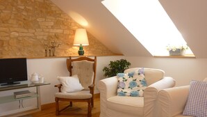 A cozy loft bedroom with a stone wall, a wooden ceiling, a TV, a glass coffee table, a wooden chair, a white sofa, and a window with a plant. - Beautiful retreat With Panoramic Countryside Views close to Hautefort Chateau. (Hautefort)