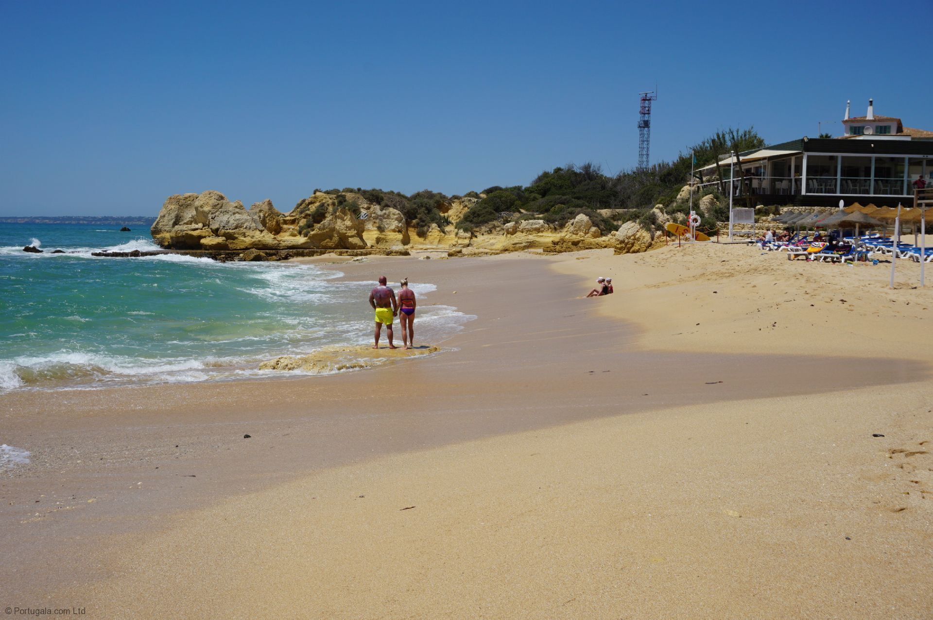 A beach with a rocky coastline, a building with a balcony overlooking the beach, and beach chairs and umbrellas.