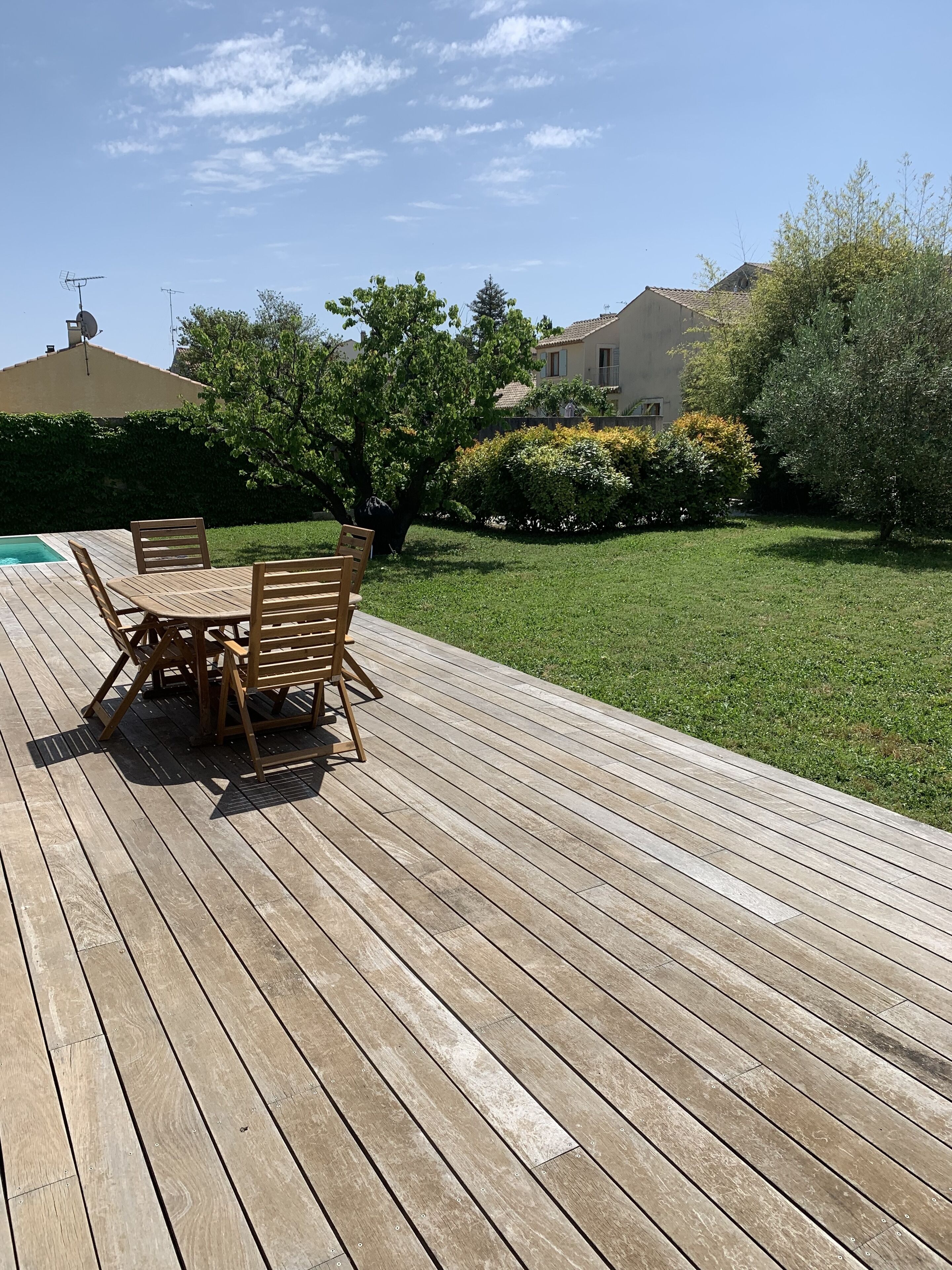 Een houten terras met een tafel en stoelen, omgeven door een verzorgd gazon en verschillende bomen.