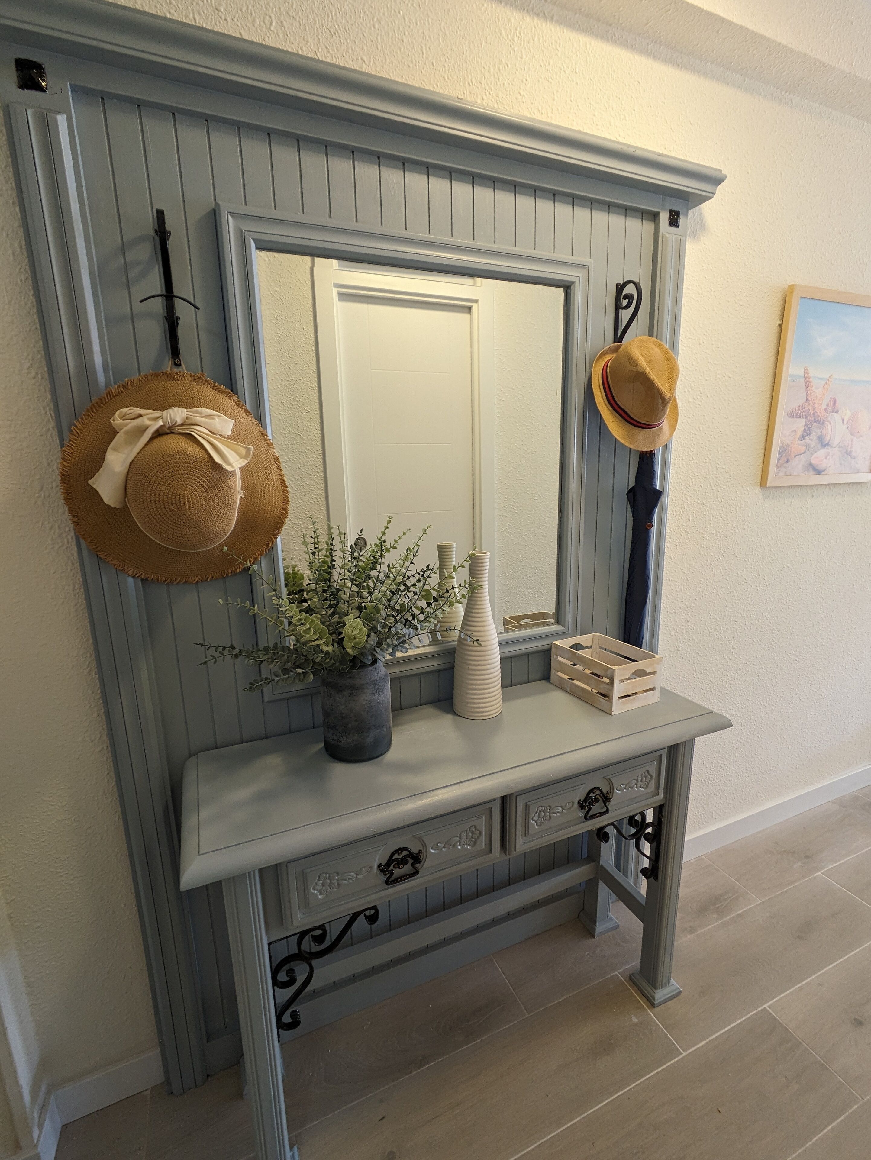 A hallway with a grey wainscoting, a mirror, a console table with decorative vases, a potted plant, and two hats hanging on hooks.