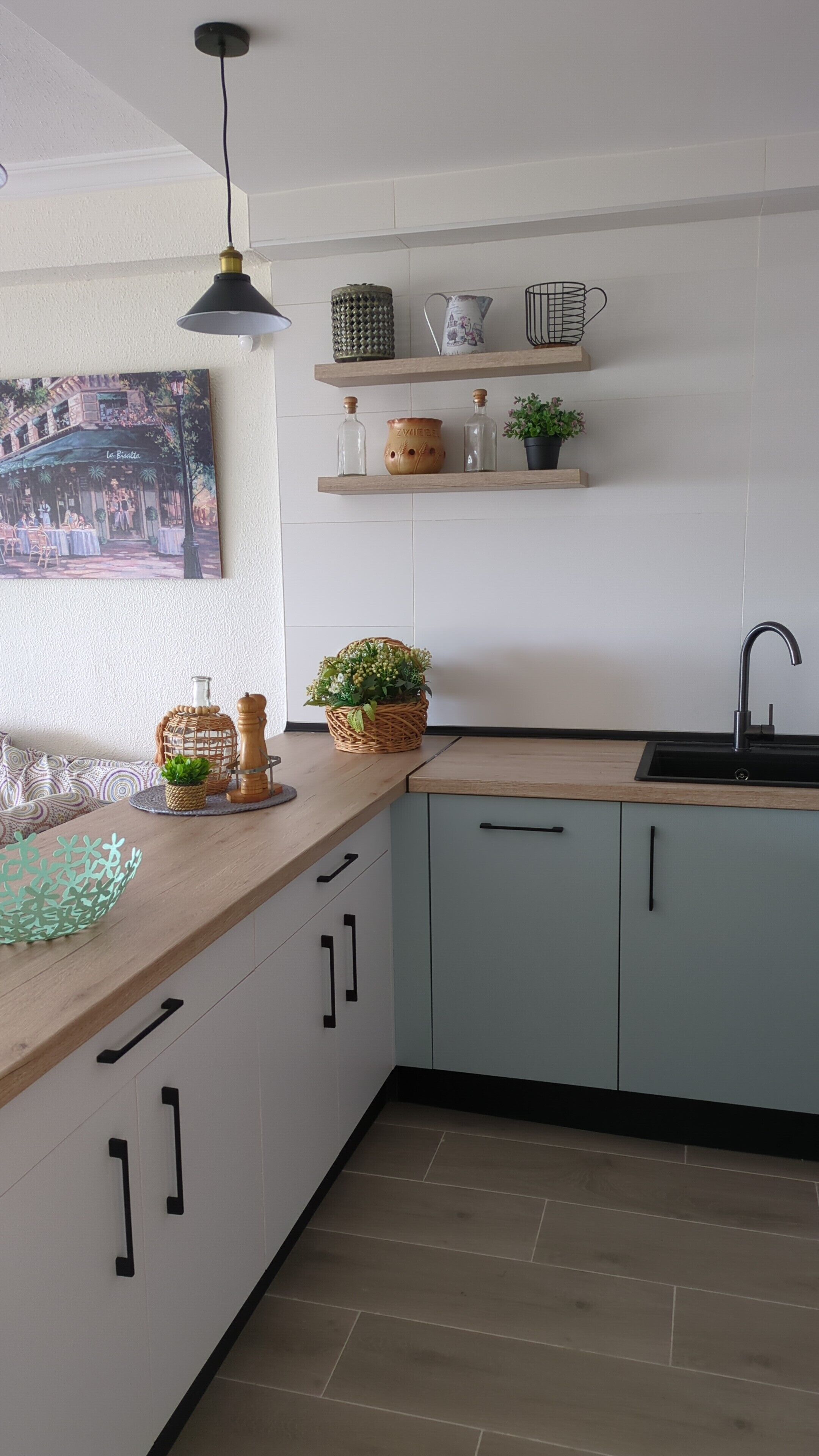 Una cocina moderna con armarios azul claro, encimera de madera y pileta negra. Hay repisas sobre la pileta con objetos decorativos y un cuadro de un café.