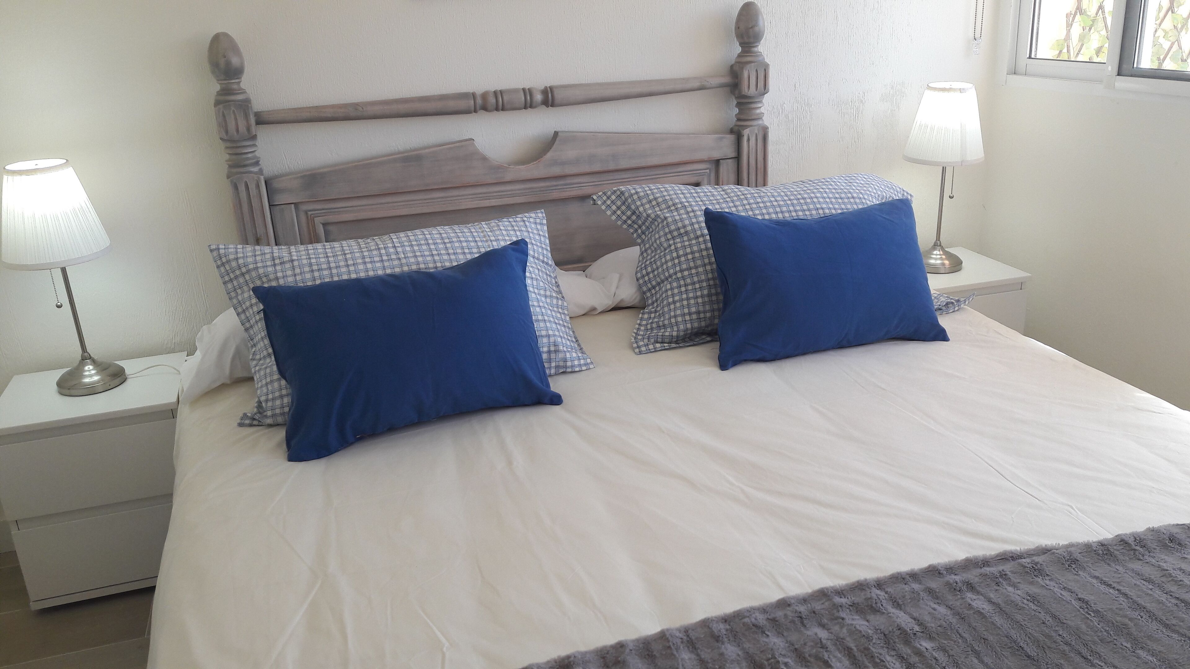 Un lit bien fait, avec une couette blanche et deux oreillers bleus, une tête de lit en bois et deux lampes de chevet posées sur des tables de chevet blanches.