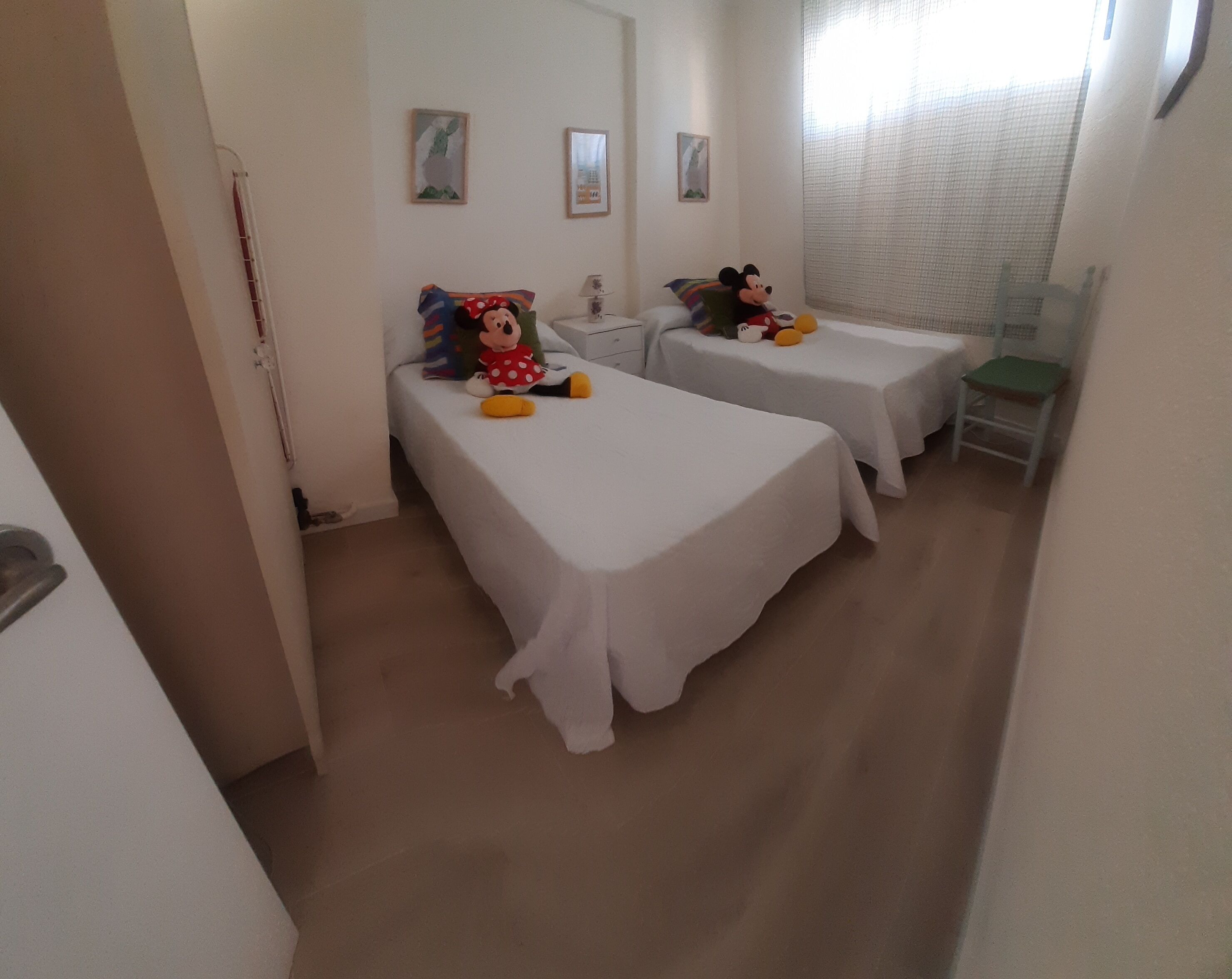 Ein Hotelzimmer mit zwei Einzelbetten, auf jedem liegt ein Mickey Mouse Kuscheltier. An der Wand hängen zwei gerahmte Bilder, ein kleiner Tisch mit einer Lampe und ein Fenster mit einem durchsichtigen Vorhang.