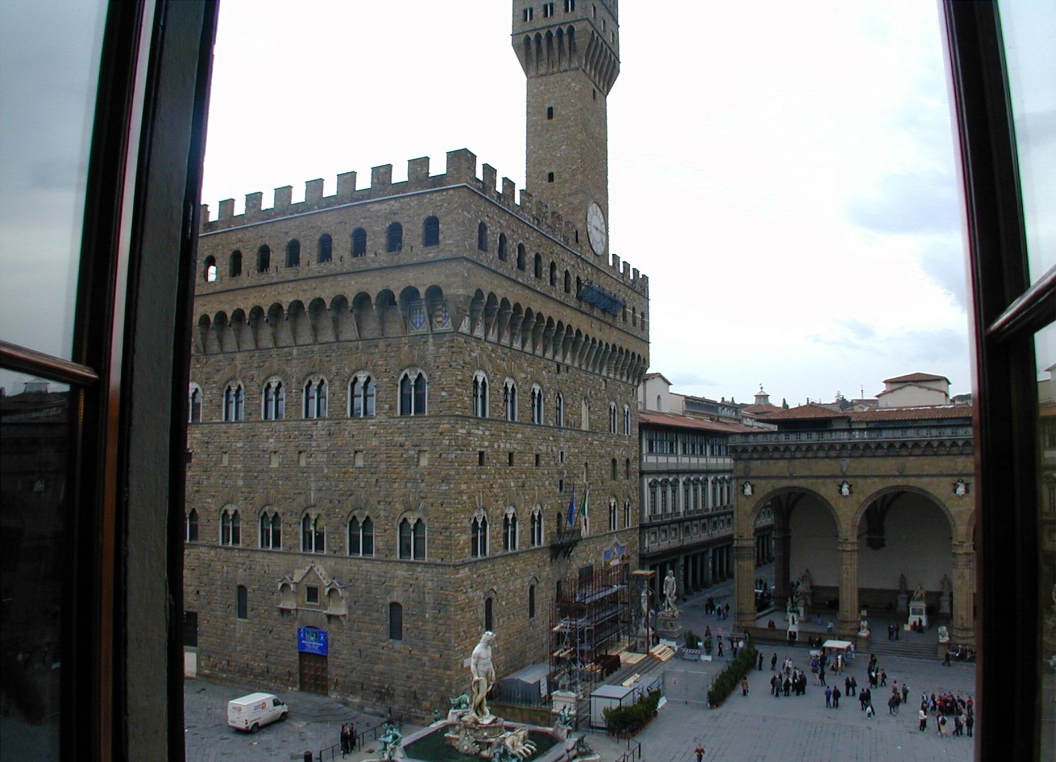 Un edificio histórico con una torre destacada y ventanas arqueadas, rodeado por una plaza con estatuas y gente.