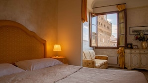 A hotel room with a large bed, a nightstand with a lamp, a chair, a window with a view of a historic building, and a cabinet with a vase of flowers. - Magic Signoria Apartment  (Firenze)