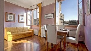 A furnished room with a wooden floor, a sofa, a dining table with chairs, and large windows offering a city view. - Magic Signoria Apartment  (Firenze)