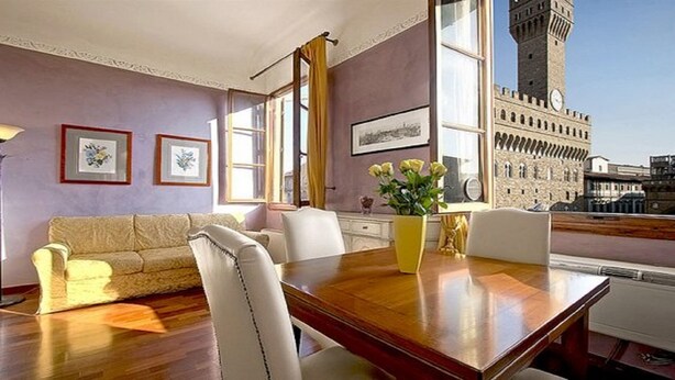 A room with a wooden dining table, white chairs, a sofa, and framed pictures on the wall. A large window offers a view of a historic building with a clock tower. - Magic Signoria Apartment  (Firenze)