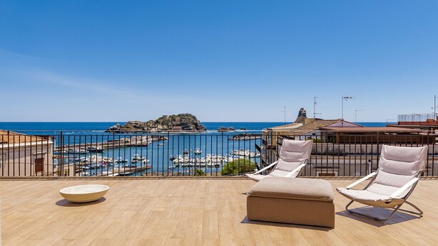 A terrace with two lounge chairs and a bowl, overlooking a marina with boats and a rocky island in the distance.