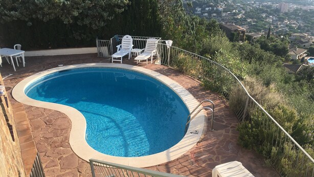 Une piscine à bord incurvé, entourée de carreaux de pierre et dotée d’une rambarde métallique. On y trouve également, à proximité, des chaises et une table en plastique blanc.