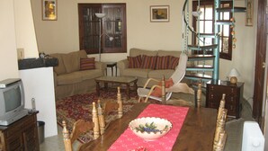 Un salon comprenant un canapé, des fauteuils, une table à manger recouverte d’une nappe rouge, un téléviseur et un escalier en colimaçon.
