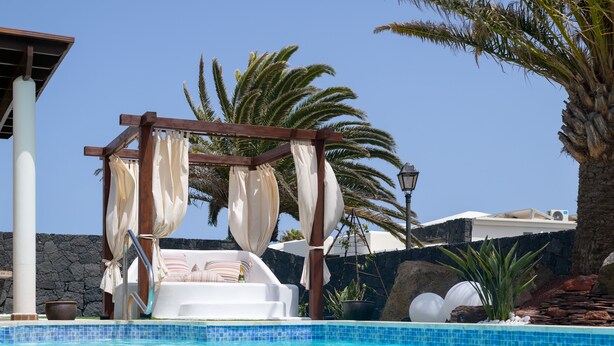 Un espace piscine agrémenté d’une chaise longue blanche, d’une pergola en bois avec des rideaux blancs et de palmiers en arrière-plan.