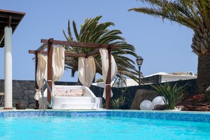 Zona de piscina con una silla lounge blanca, una pérgola de madera con cortinas blancas y palmeras al fondo.